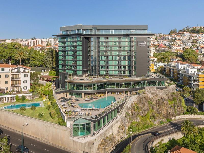Modernes Glasgebäude auf Felsen mit Pool, umgeben von Wohnhäusern unter klarem Himmel.