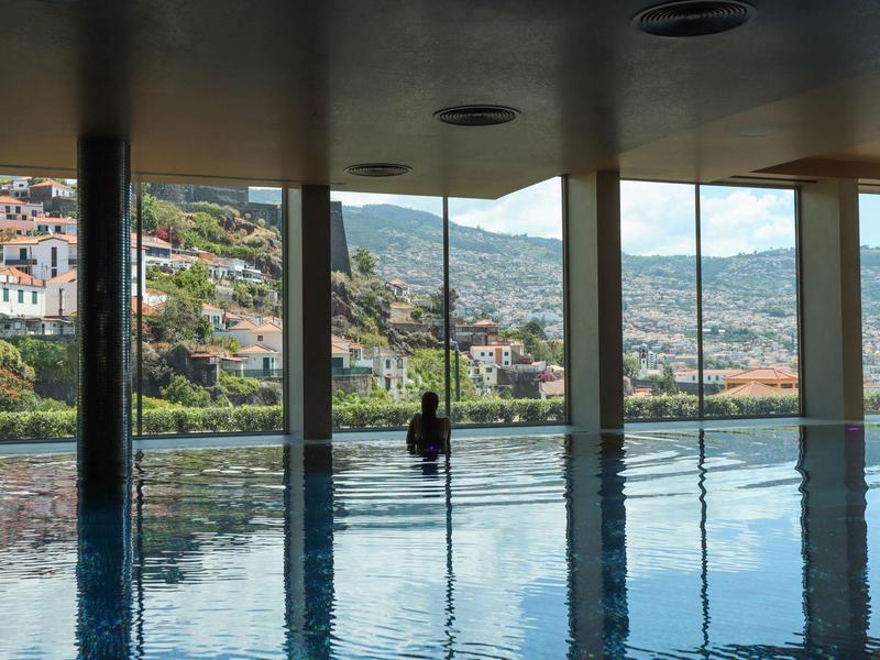Ein Mann sitzt am Rand eines Pools mit Blick auf eine hügelige Stadtlandschaft durch große Fenster.