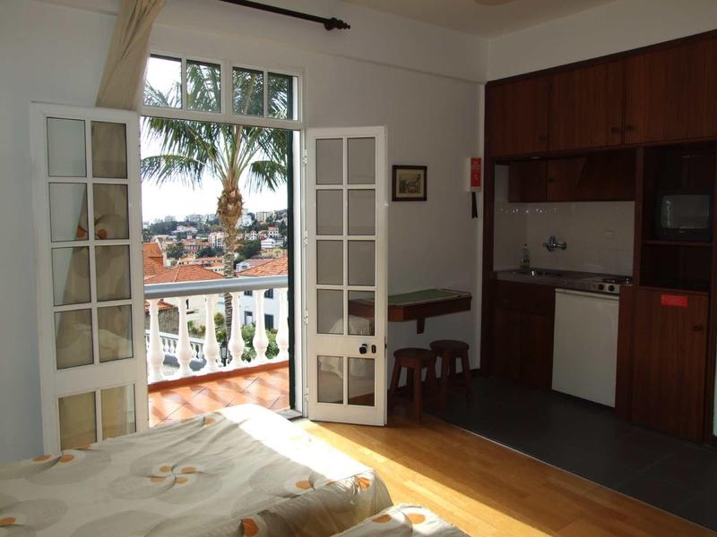 Helles Zimmer mit offener Tür zum Balkon, Holzmöbeln und kleiner Küchenzeile.