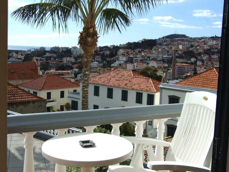 Balkon mit weißem Tisch und Stuhl, Palmenblatt, rote Dächer und Stadt im Hintergrund.
