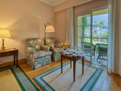 Soggiorno accogliente con poltrona, tavolo e balcone con vista su montagne e giardino.