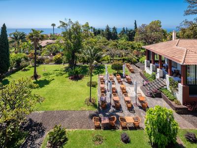 Hotelová terasa s stoly a židlemi vedle dobře udržované zahrady s stromy a výhledem na moře.