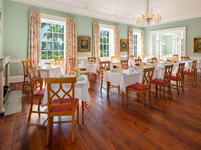 Sala da pranzo luminosa con pavimento in legno, più tavoli con tovaglie bianche e sedie in legno.