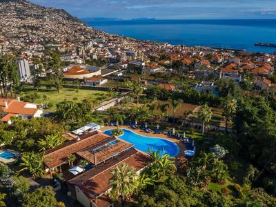 Vista aerea di un hotel con piscina, circondato dalla città e dal mare sullo sfondo.