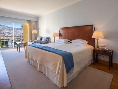 Camera d'hotel luminosa con letto grande, mobili in legno e balcone con vista sulle montagne.