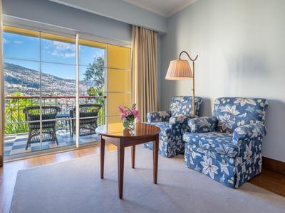 Sala d'hotel luminosa con due poltrone a motivo, tavolo in legno e balcone con vista mare.