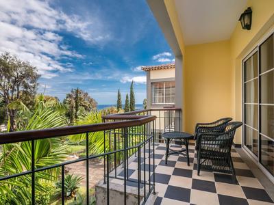 Balcone con ringhiera in metallo nero, pavimento piastrellato, sedie e tavolo, vista sul giardino e sul cielo.