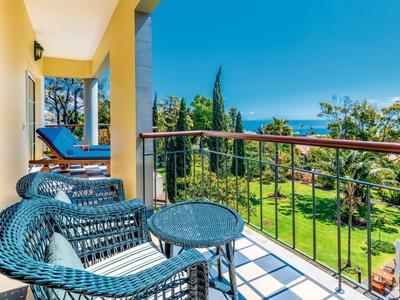 Balcone con sedie in vimini che si affaccia su un giardino verde e sul mare sotto un cielo limpido.