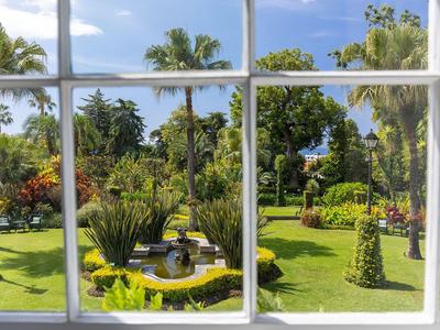 Blick durch ein Fenster auf einen sonnigen, gepflegten Hotelgarten mit Palmen und Springbrunnen.