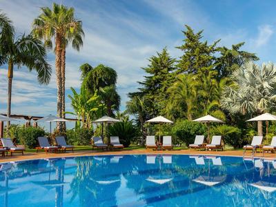 Ein Swimmingpool mit Liegestühlen und Sonnenschirmen in einer tropischen Gartenanlage bei blauem Himmel.
