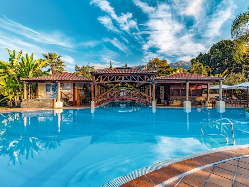 Grande piscina all'aperto con ponte davanti a un hotel tropicale sotto il cielo azzurro.