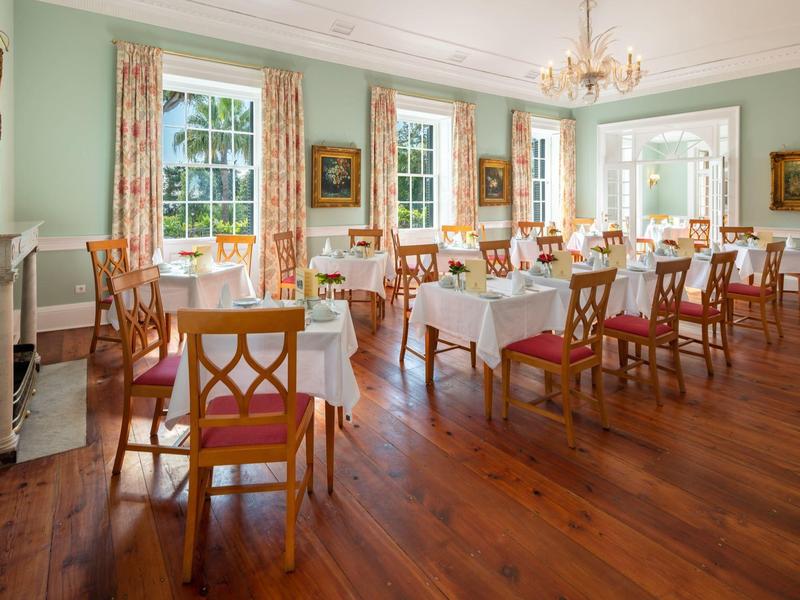 Sala da pranzo luminosa con pavimento in legno, più tavoli con tovaglie bianche e sedie in legno.