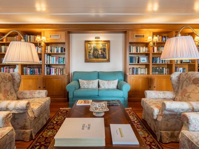 Accogliente sala lettura con scaffali, tappeto, divano e poltrone in luce calda.