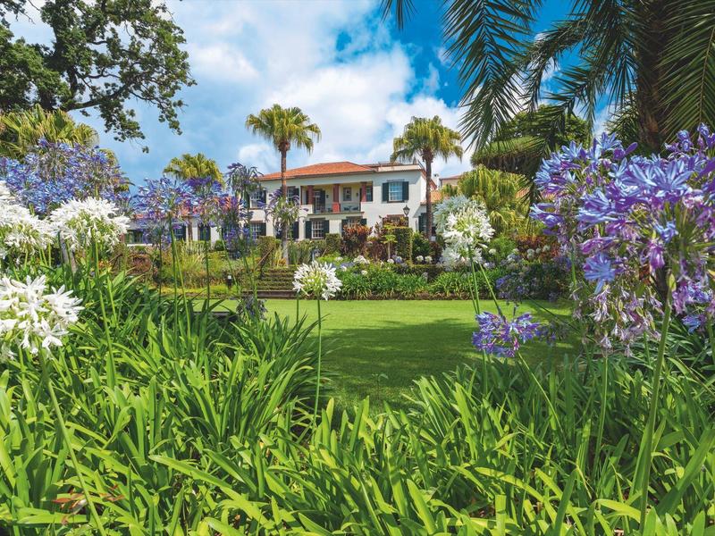 Un edificio dell'hotel dietro un giardino fiorito con palme e prato verde sotto un cielo blu.