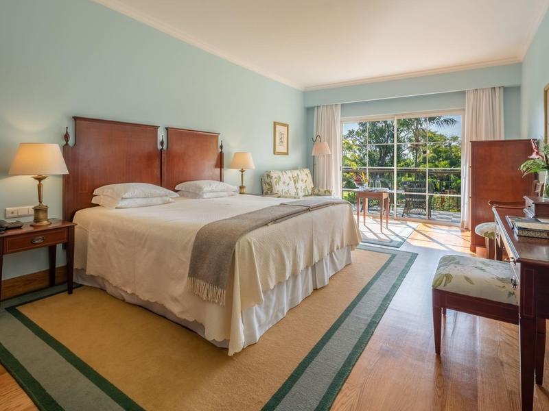 Camera d'hotel luminosa con letto matrimoniale, scrivania, lampada da terra e balcone con vista sul verde.