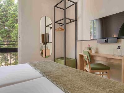 Chambre d'hôtel lumineuse avec deux lits, bureau, chaise et fenêtre donnant sur la verdure.