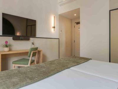 Espace de chambre d'hôtel moderne avec lit, bureau, chaise et télévision murale en tons neutres.