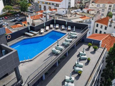 Terrasse d'hôtel avec piscine, chaises longues et bâtiments environnants par une journée ensoleillée.