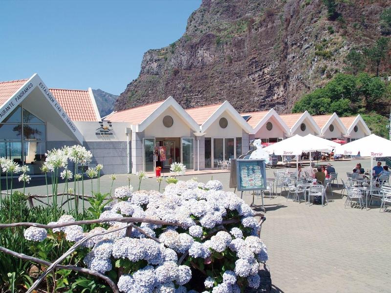 Small hotel with white gardens and seating area in front of a rocky mountain under clear sky.