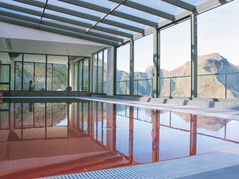 Indoor pool with glass walls offers view of mountain landscape on cloudy day.