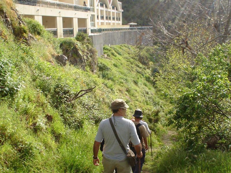 Hikers on a green mountain trail with small buildings in the background.