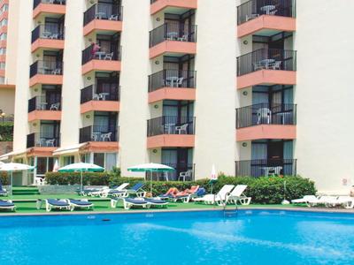 Hotel mit mehreren Stockwerken, Balkonen und Poolbereich mit Liegestühlen und Sonnenschirmen.