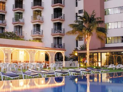 Piscine d'hôtel éclairée avec chaises longues et palmiers devant un immeuble de plusieurs étages au crépuscule
