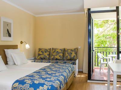 Lichte hotelkamer met tweepersoonsbed, patroonbedsprei en toegang tot balkon met uitzicht op groene bomen.