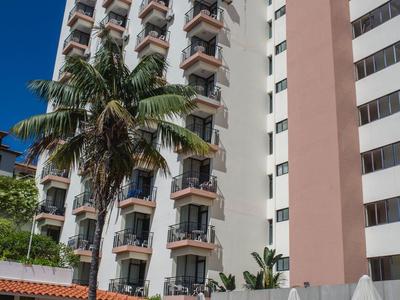 Hoog hotelgebouw met veel balkons en een zwembadgedeelte met palmbomen en parasols.
