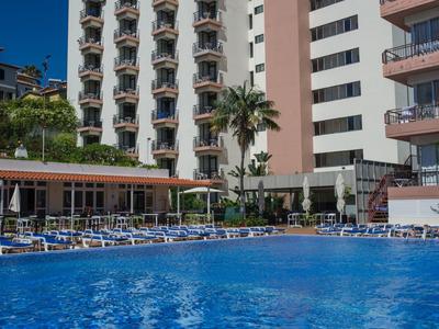 Hotelgebouw met meerdere balkons, zwembad en ligstoelen onder een heldere lucht.