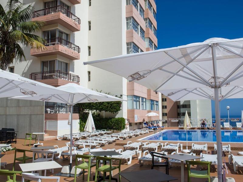 Buitenzwembad van een hotel met parasols, tafels en stoelen bij heldere lucht.