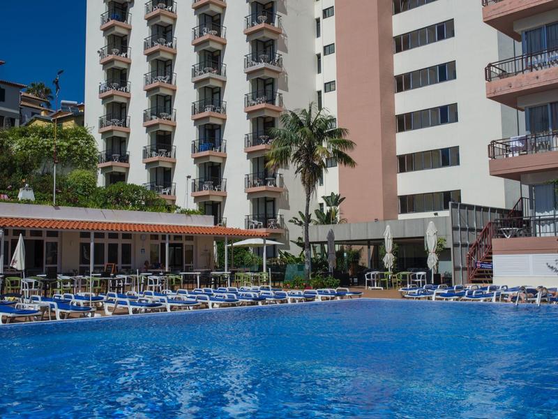 Hotelgebouw met meerdere balkons, zwembad en ligstoelen onder een heldere lucht.