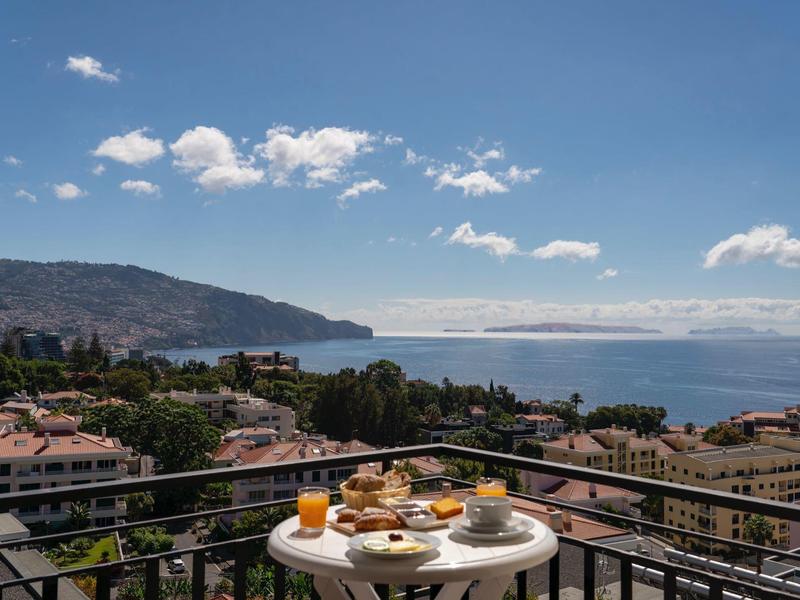 Balkon met ontbijttafel met uitzicht op stad en zee onder een blauwe lucht.
