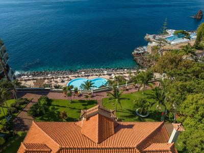 Blick auf Hotel mit Pool, Palmen, grüner Wiese und Meer im Hintergrund an sonnigem Tag.