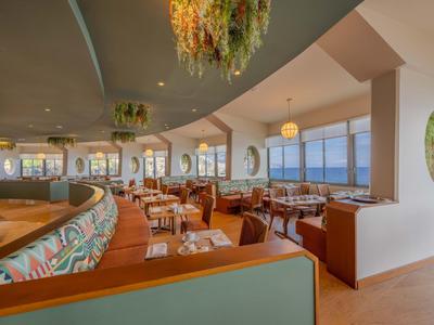 Modernes Restaurant mit großen Fenstern, Holzvertäfelungen und grünen Sitzpolstern am Meer.