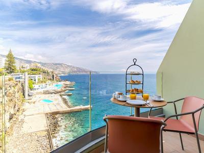 Balkon mit Tisch, Stühlen und Frühstück, Meerblick und Felsen entlang der Küste bei klarem Himmel.