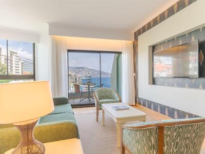 Helles Wohnzimmer mit großen Fenstern, grünem Sofa, Sesseln und Blick auf das Meer.