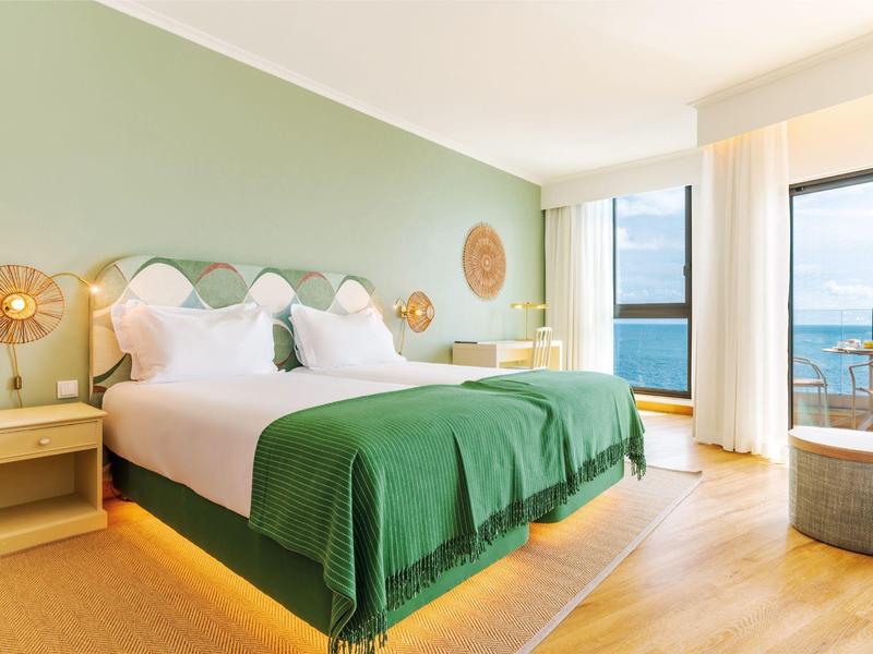 Helles Hotelzimmer mit Doppelbett, grünen Decken, Holzboden und Meeresblick durch großes Fenster.