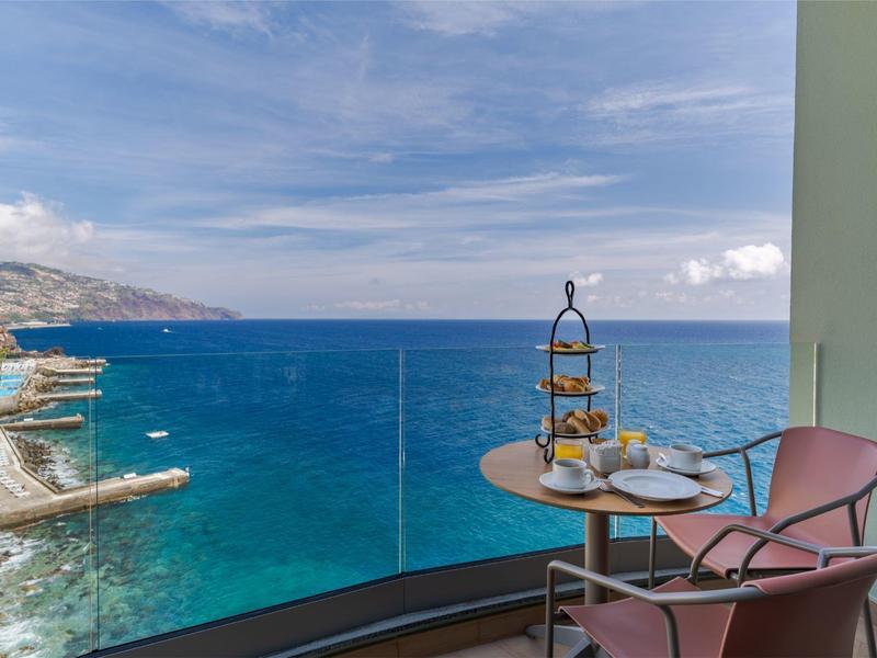 Balkon mit Tisch, Stuhl und Snacks mit Blick auf das blaue Meer und bewölkten Himmel.