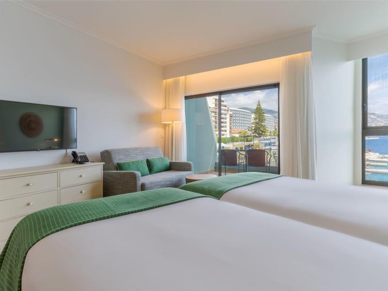 Modernes Hotelzimmer mit Twin-Betten, grüner Couch und Meerblick durch große Fenster.