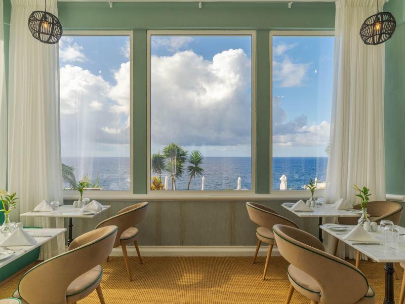 Helles Restaurant mit Panoramafenstern und Blick auf das Meer und Palmen.