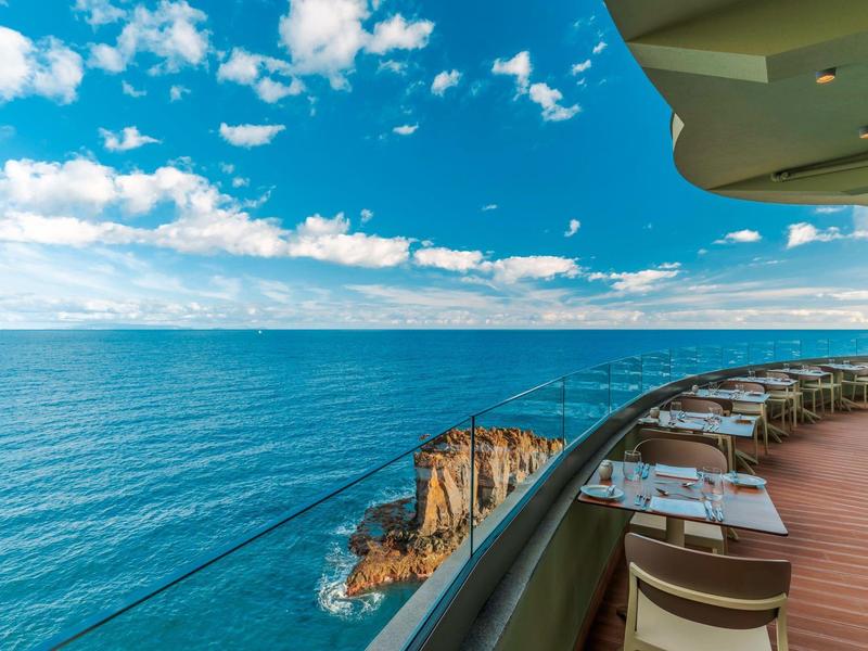 Balkonrestaurant mit Meerblick und Felsen unter blauem Himmel.