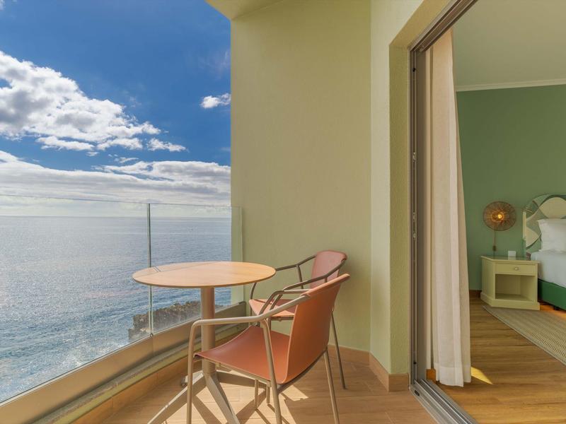 Balkon mit Tisch und zwei Stühlen mit Blick aufs Meer bei klarem Himmel.