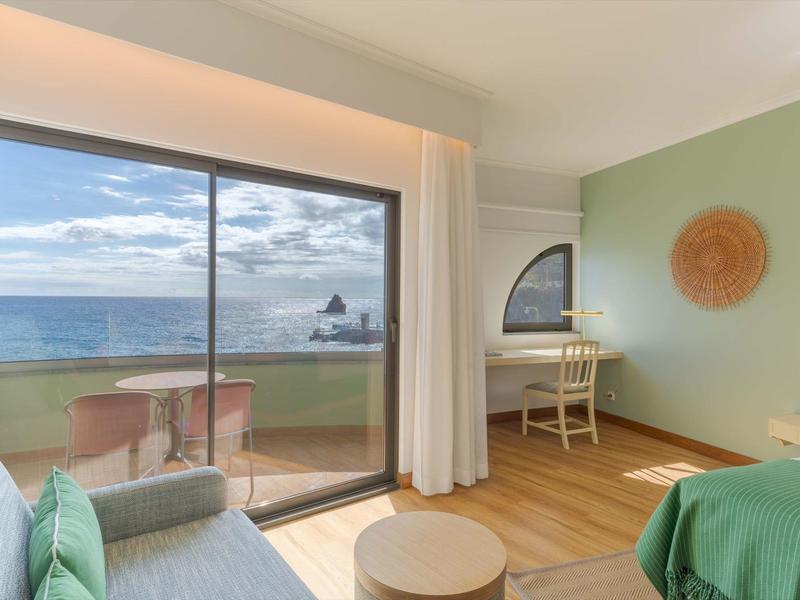 Helles Zimmer mit Bett, Sofa, Tisch und Meerblick durch große Glasschiebetür.