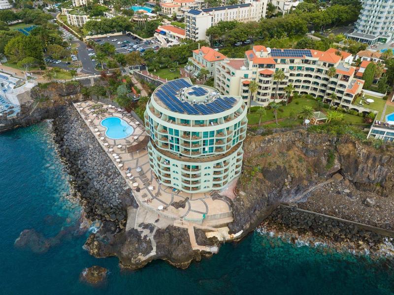 Rundes Hotel mit Pool auf Klippe am Meer, umgeben von weiteren Gebäuden und Grünflächen.
