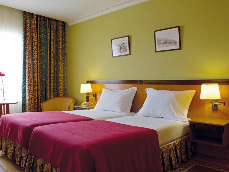 Helles Hotelzimmer mit zwei Einzelbetten, roten Bettdecken und gelben Wänden.