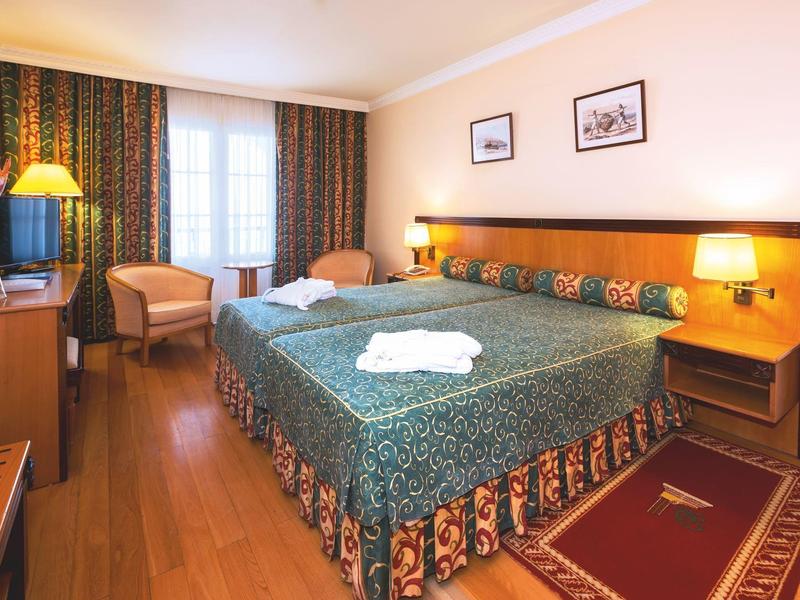 Ein geräumiges Hotelzimmer mit Doppelbett, Teppich, Sesseln, Nachttischlampen und Fenster mit Vorhängen.