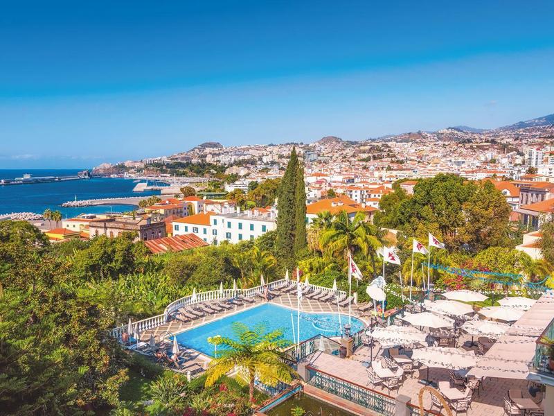 Blick auf einen Hotelpool mit Liegen und Stadt am Meer im Hintergrund an einem sonnigen Tag.