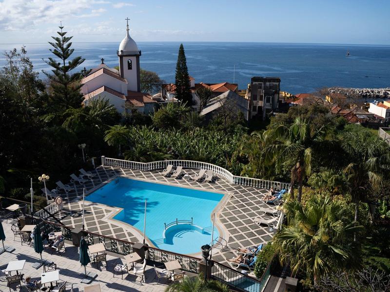 Blauer Pool mit Liegestühlen, umgeben von Palmen und Gebäuden, dahinter Meer und Himmel.