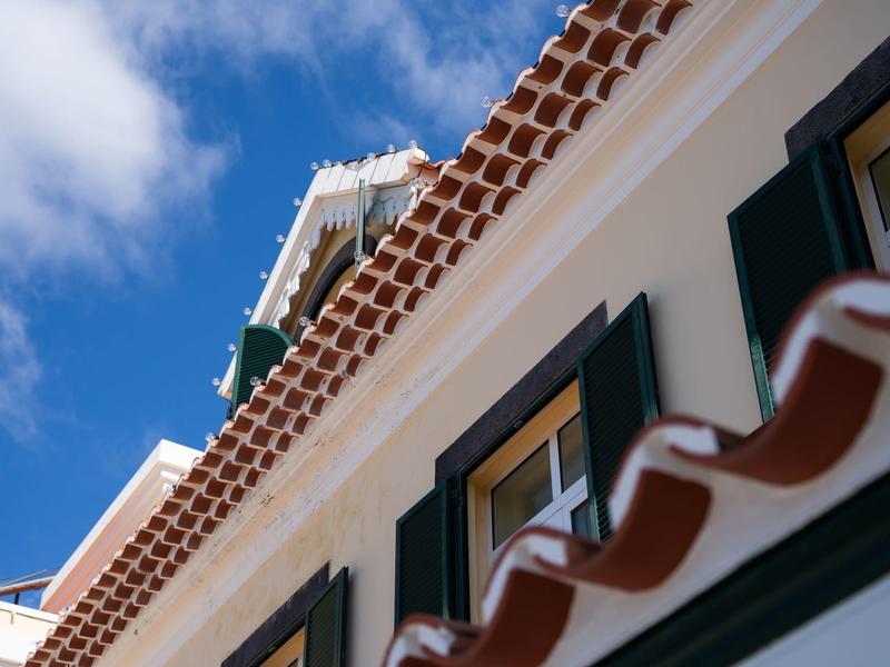 Schräger Blick auf ein weißes Haus mit roten Ziegeldächern und grünen Fensterläden bei blauem Himmel.
